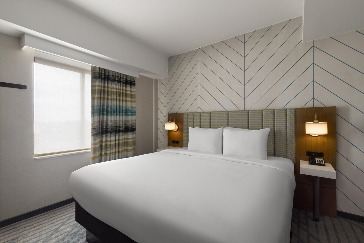 A hotel room with a large bed, two bedside tables with lamps, a window with curtains, and a chevron-patterned wall.