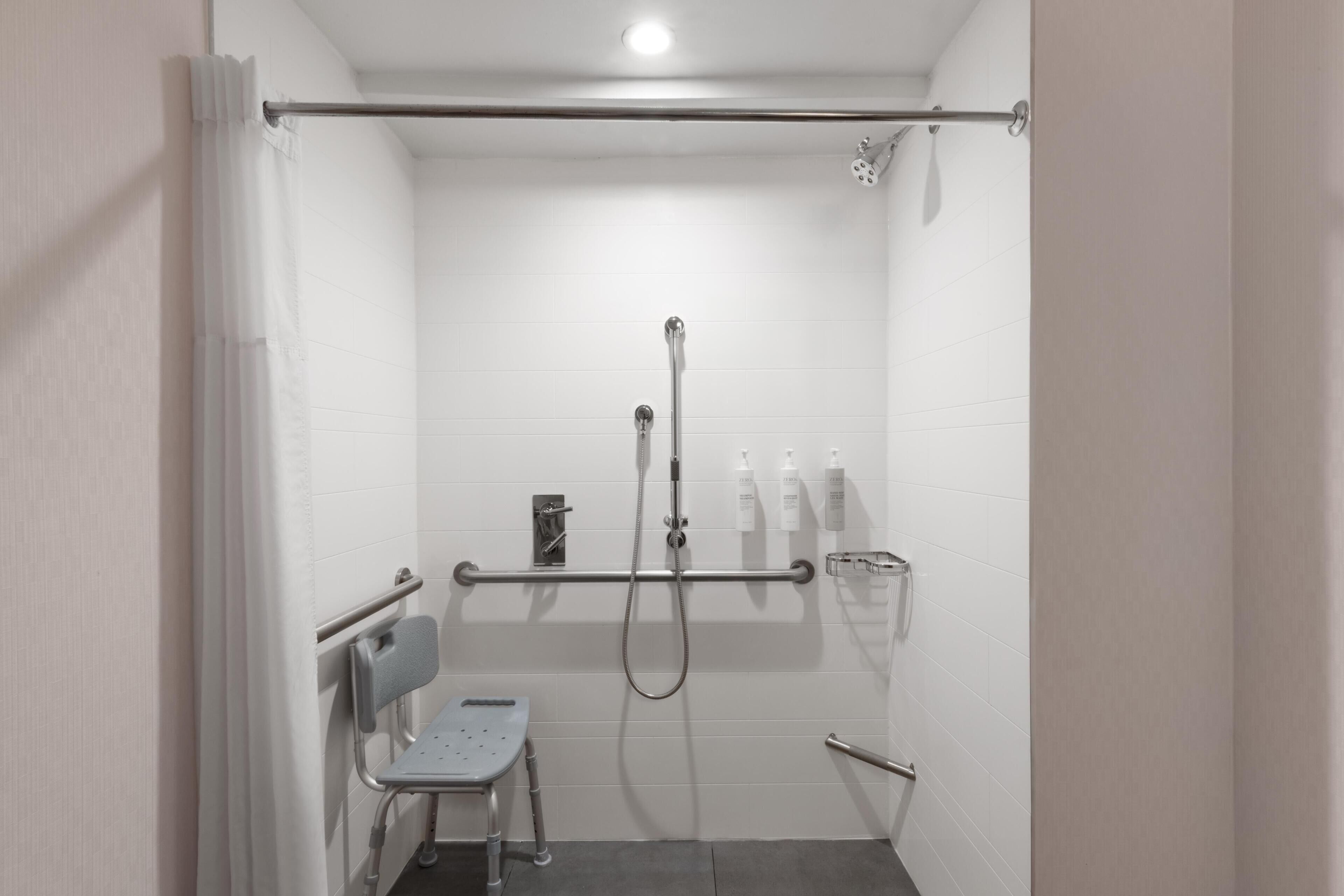 Un baño pequeño con paredes blancas, ducha, barra de apoyo, banco accesible para sillas de ruedas y pasamanos.