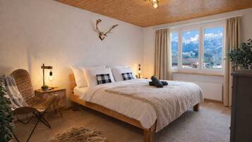 Ein Schlafzimmer mit Holzboden, einem Bett mit weißer Bettwäsche, einem aufgehängten Damhirschschädel, einem Fenster mit Vorhängen und Blick ins Freie.
