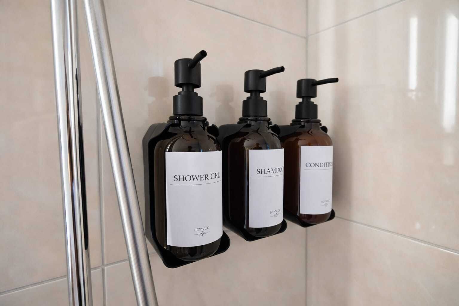 Trois bouteilles de gel douche, de shampoing et d’après-shampoing sont posées sur un mur carrelé.
