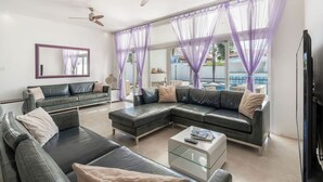 Ein Wohnzimmer mit einer Eckcouch, einem Glastisch, einem Deckenventilator und großen Fenstern mit violetten Vorhängen.