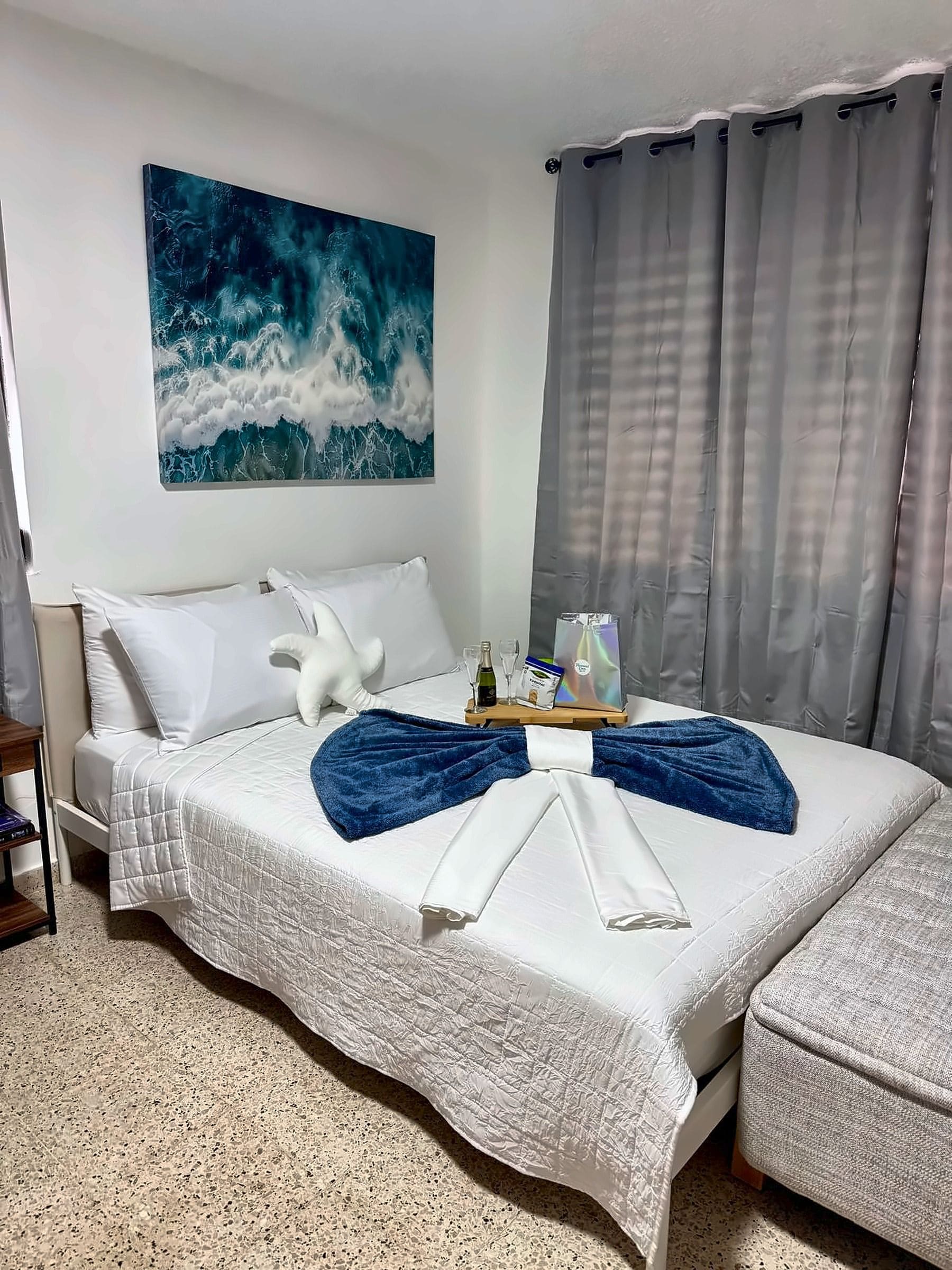 A hotel room with a bed, a bedside table, a painting, and a window with curtains.