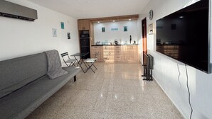 Een knus woongedeelte met een kitchenette, een grijze bank en een flatscreen televisie aan de muur.