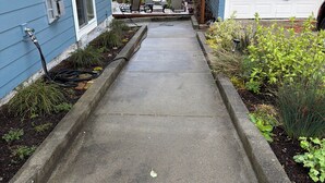 Un chemin en béton mène à un espace patio aménagé avec du mobilier d’extérieur et un garage.