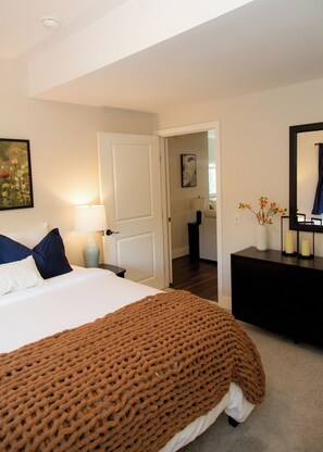 A bedroom with a bed, a nightstand, a dresser, a mirror, and a door leading to another room. - Garden-Level 2BR Suite, West Seattle (Seattle)