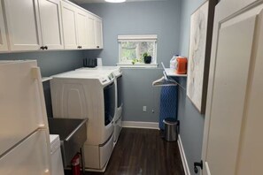 A compact laundry room with a washer and dryer, a window with a view, and a small shelf with cleaning supplies. - Garden-Level 2BR Suite, West Seattle (Seattle)