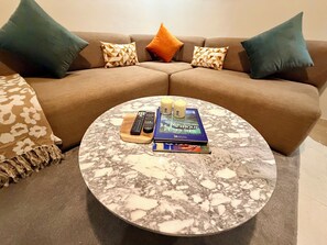 Ein Wohnzimmer mit einem braunen Sofa, zwei Kissen, einem Couchtisch mit Büchern und Kerzen sowie Fernbedienungen.