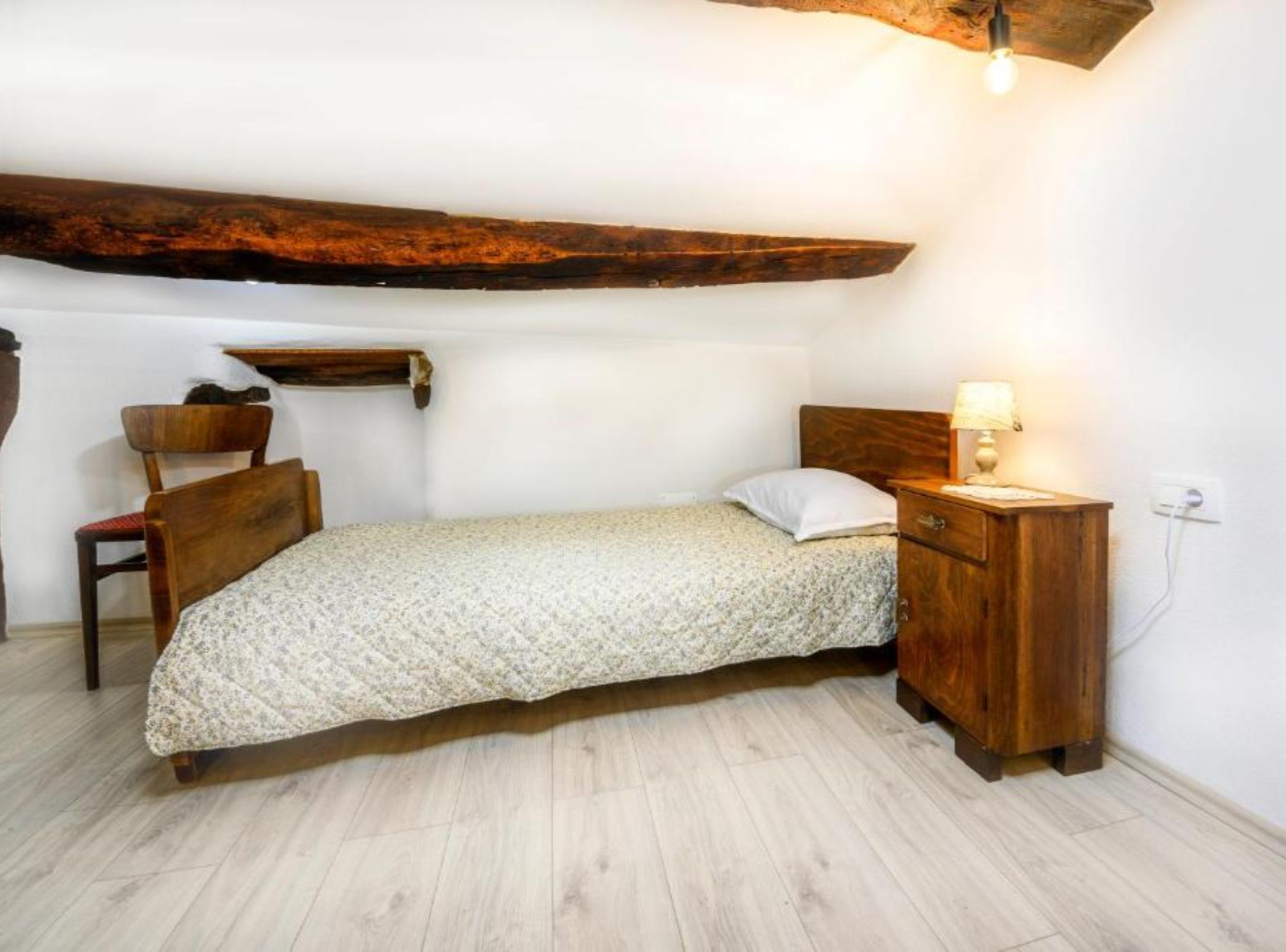 A small, cozy bedroom with a single bed, a wooden nightstand, a chair, and a shelf.