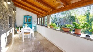 Una habitación con pared de piedra, vigas de madera en el techo, una mesa con sillas, un armario azul y plantas en macetas junto a una ventana.