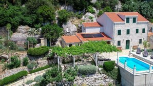 A stone building with a red-tiled roof, a swimming pool, and a garden with various plants and a pergola.
