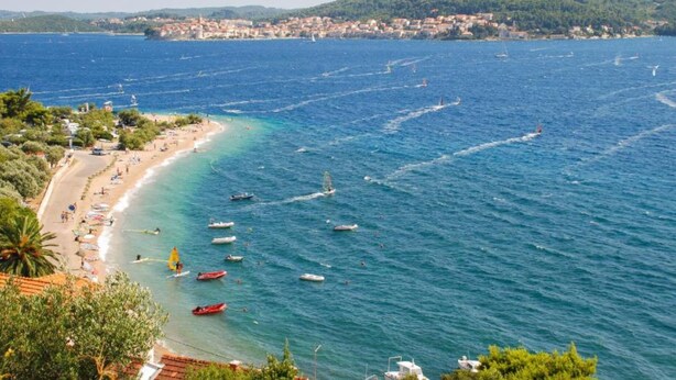 A coastal beach with clear waters, boats, and a distant town.
