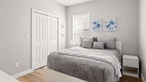 Ein Schlafzimmer mit einem Bett, Nachttischen, einem Fenster mit Jalousien und zwei Blumenbildern an der Wand.