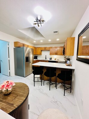 A modern kitchen with wooden cabinets, a central island with bar stools, a refrigerator, and a ceiling light fixture. - 3-bedroom house in gorgeous Burleson with AC for relaxing stays (Burleson)