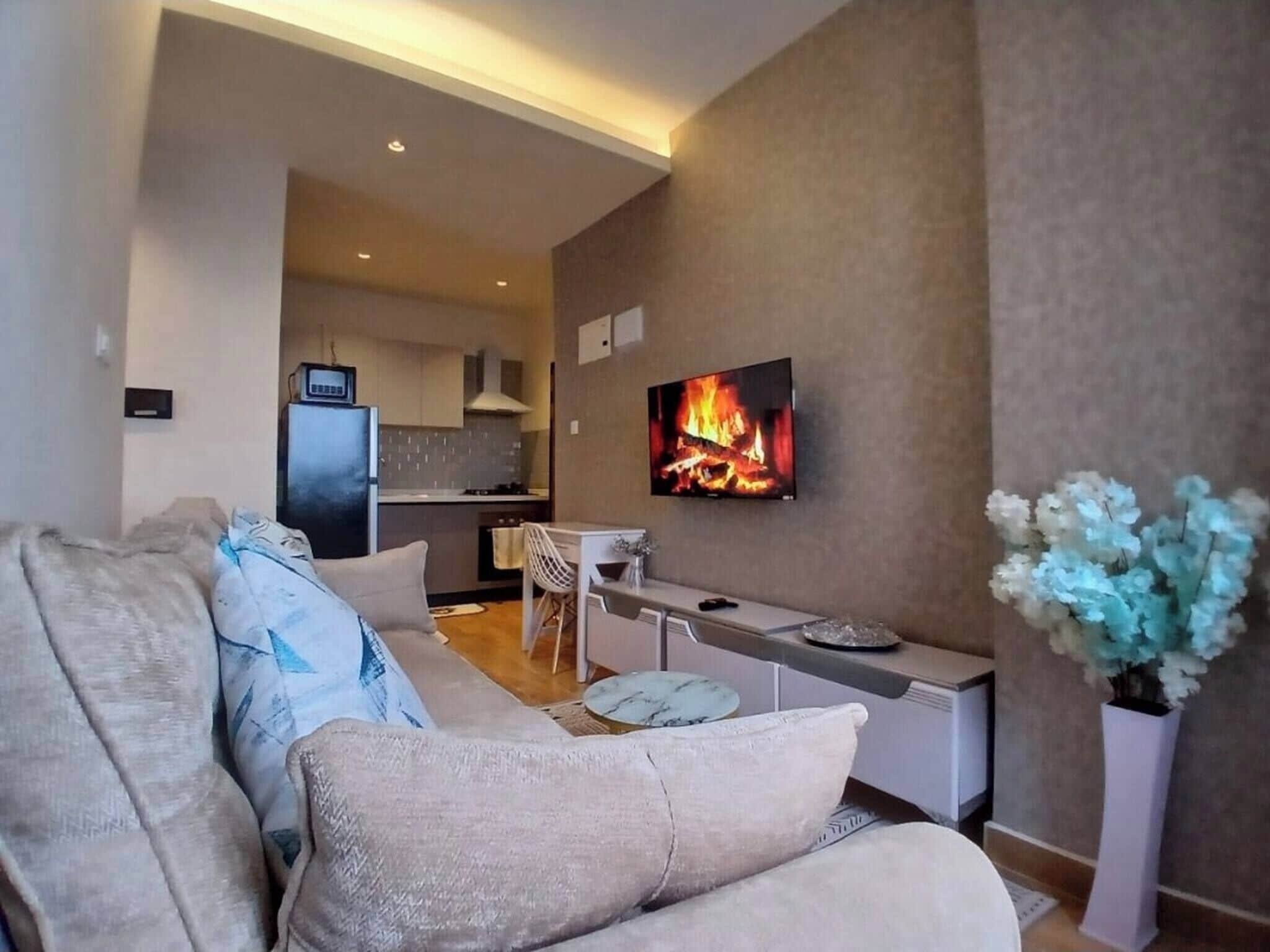 A modern living room with a sofa, a TV displaying a fireplace, a kitchen in the background, and a vase with flowers.