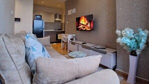 A modern living room with a sofa, a TV displaying a fireplace, a kitchen in the background, and a vase with flowers. - Executive one Bedroom Kilimani (Nairobi)