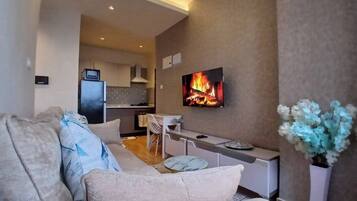 A modern living room with a sofa, a TV displaying a fireplace, a kitchen in the background, and a vase with flowers.