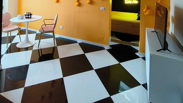 A room with a checkered floor, a wall with a pattern of orange shapes, a neon sign, and a television. - The Rabbit Hole: Unique Design Stay in Milan Downtown with AC and Wifi (Milano)
