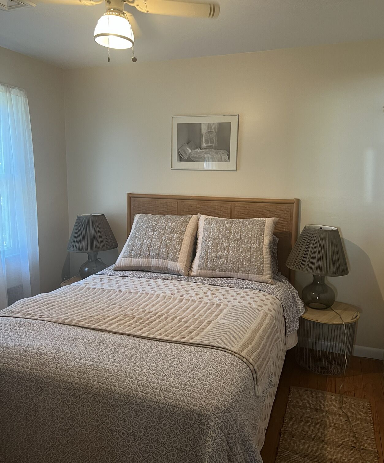 A bedroom with a bed, two bedside lamps, a ceiling fan, a framed picture on the wall, and a window with curtains.