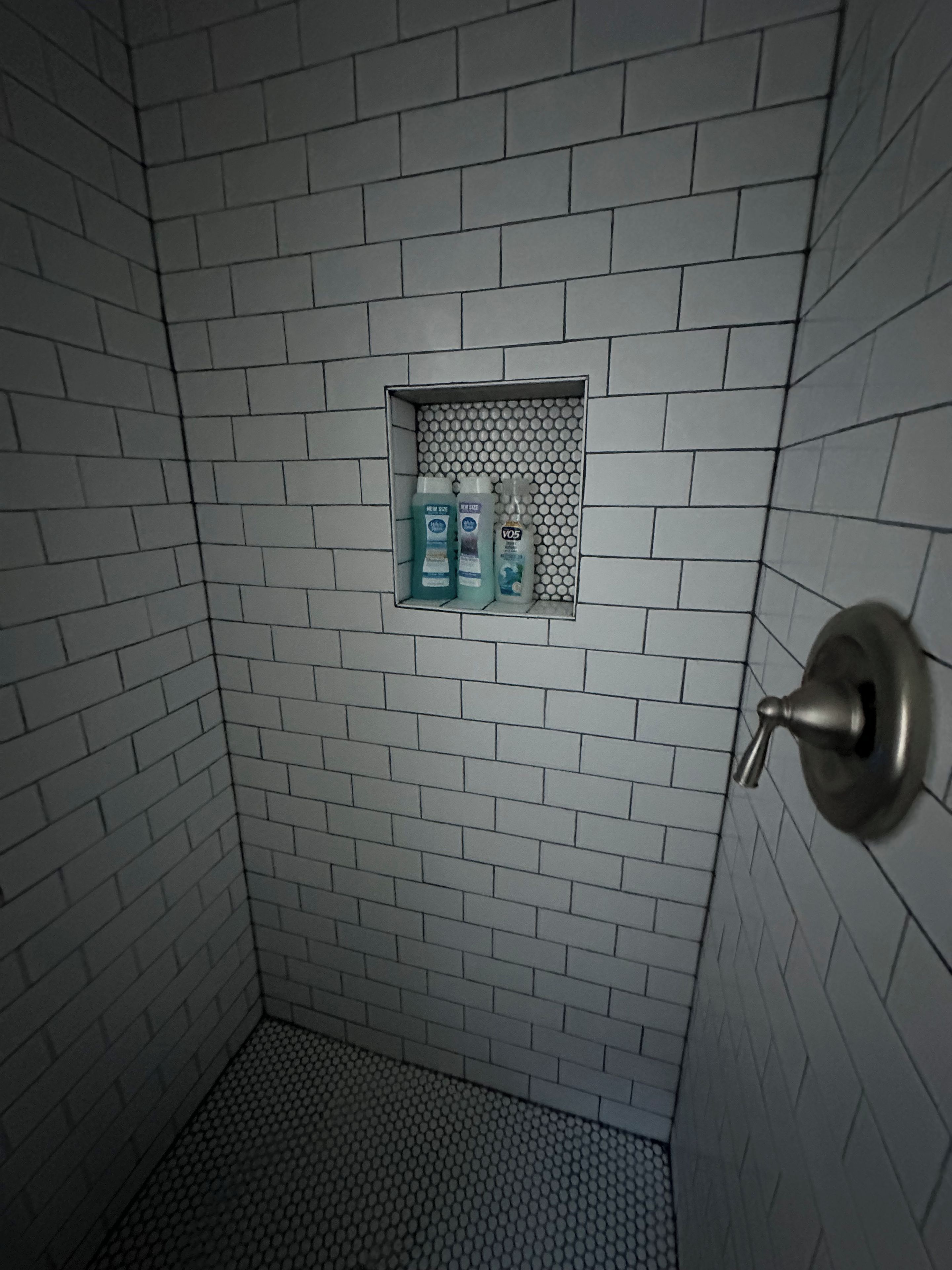 A bathroom with white tiled walls, a shower area, and a shelf with toiletries.