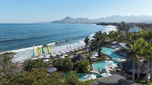 A resort with a pool area, beach umbrellas, and a view of the ocean and mountains. - Spectacular Kupuri Beach Oceanfront 6BR Luxury Villa, Infinity Pool, Chef (Punta Mita)