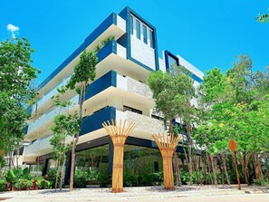 A modern multi-story building with a flat roof, surrounded by lush greenery and bamboo structures. - Centric 2BD Condo in Aldea Zama (Tulum)