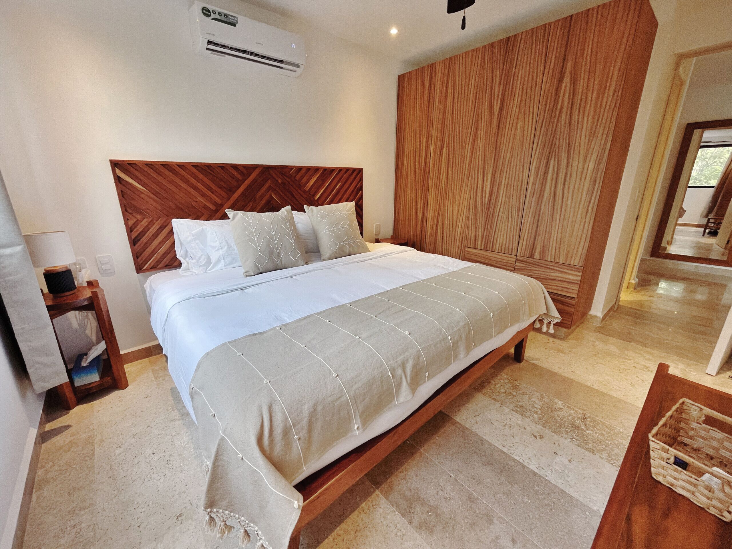 A bedroom with a wooden headboard, a bed with white bedding, a nightstand, an air conditioning unit, and a wooden wardrobe.