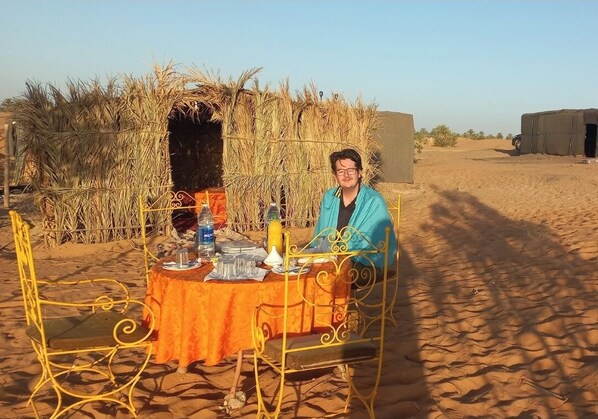 A person seated at a table with a tablecloth, surrounded by metal chairs, in a desert setting with sparse vegetation and a clear sky.