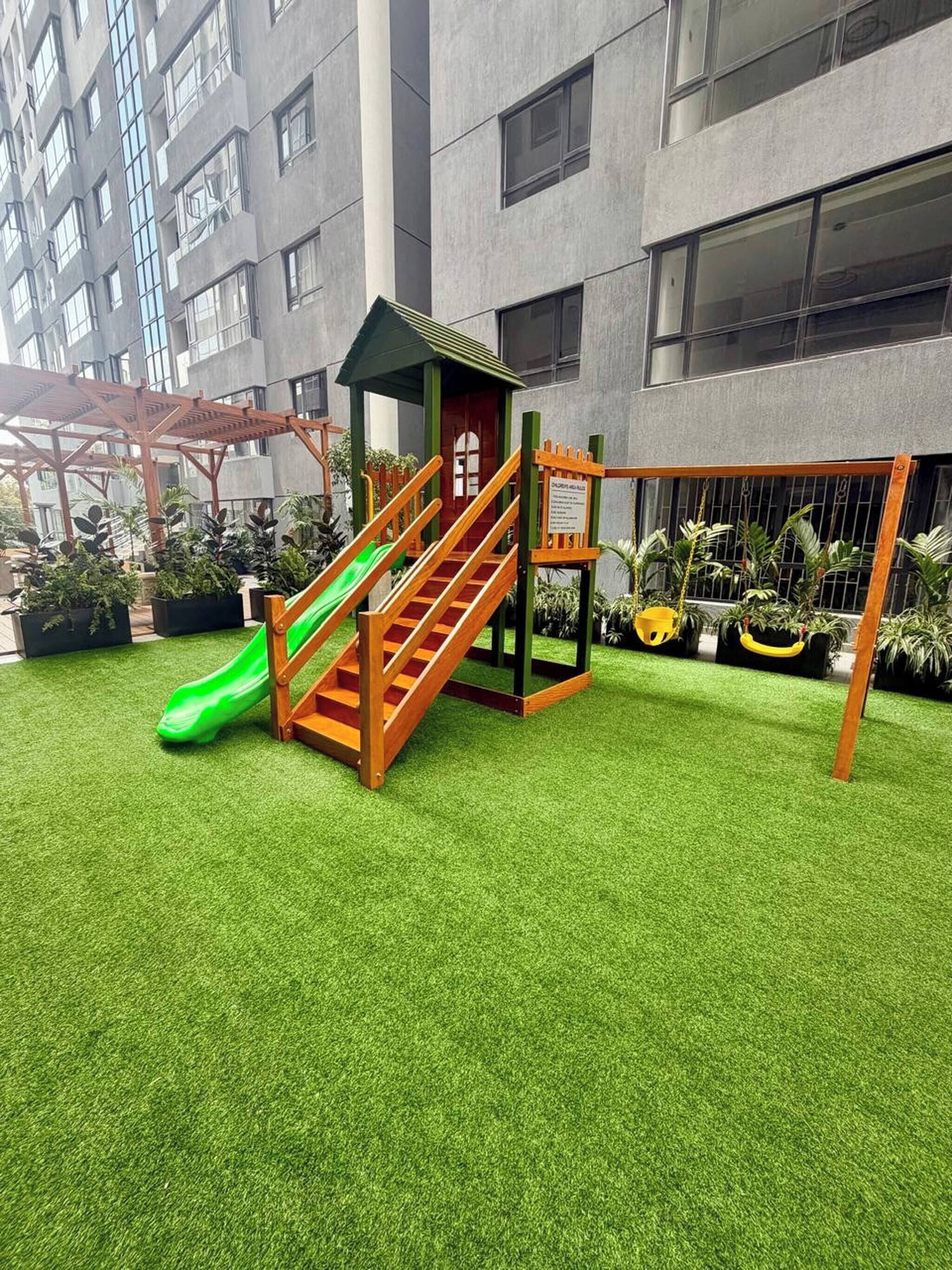 Uma área de recreação infantil com escorregador e estrutura de escalada, cercada por grama sintética, em frente a um edifício moderno com amplas janelas.