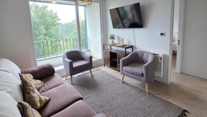 Een moderne woonkamer met een bruine hoekbank, twee fauteuils, een kamerbreed tapijt, een aan de muur bevestigde televisie, en een balkon met uitzicht op groen.