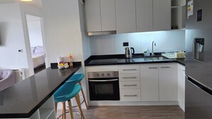 Een moderne keuken met witte kasten, een zwart aanrecht en een inbouwoven. Er zijn twee blauwe barkrukken en een hangende lamp.