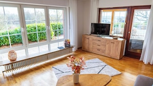 Uma sala de estar com piso de madeira, um sofá, uma mesa de centro, uma TV sobre um móvel de madeira e uma janela com vista para o verde.