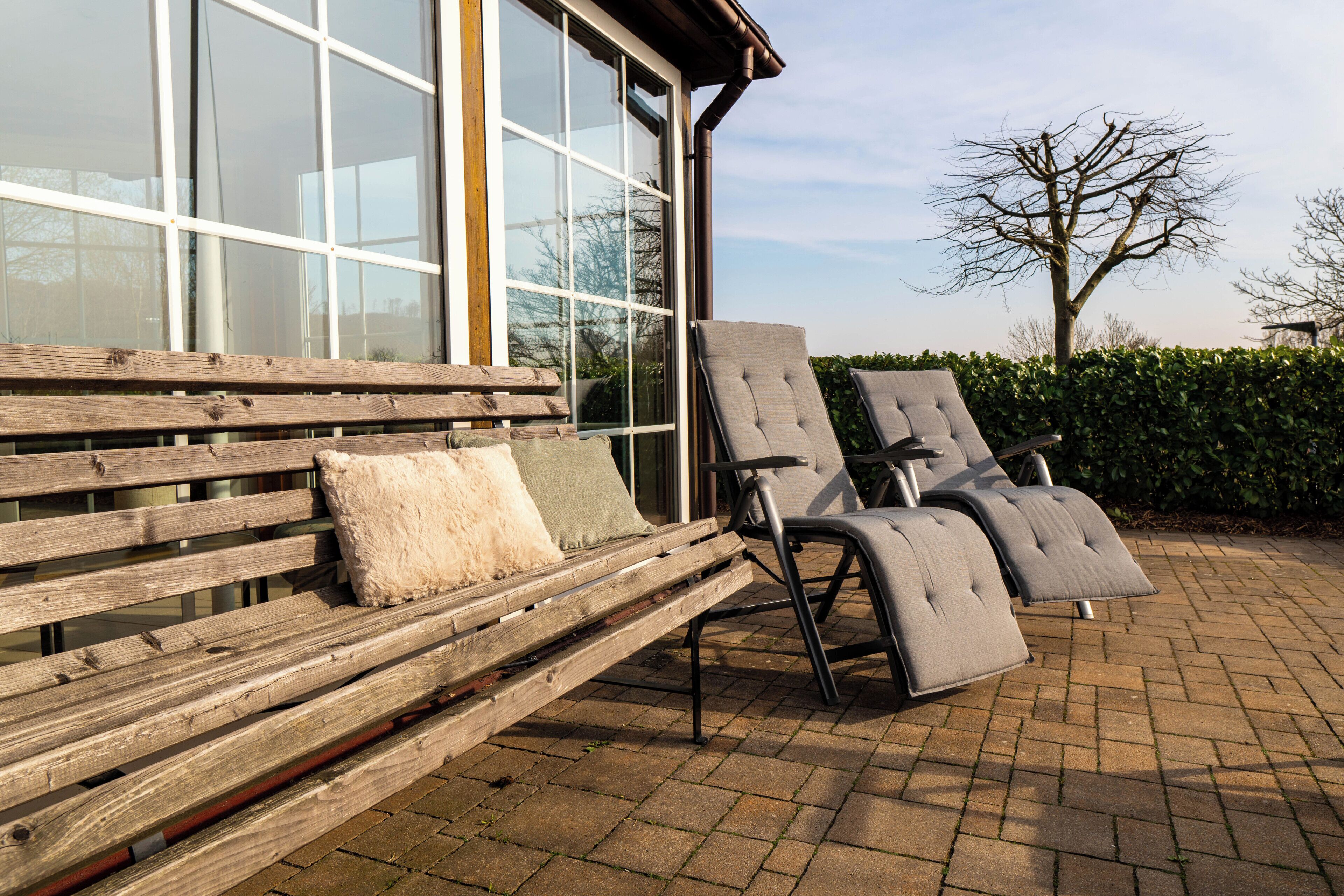 A wooden bench with cushions and two grey lounge chairs outside a building with large windows.