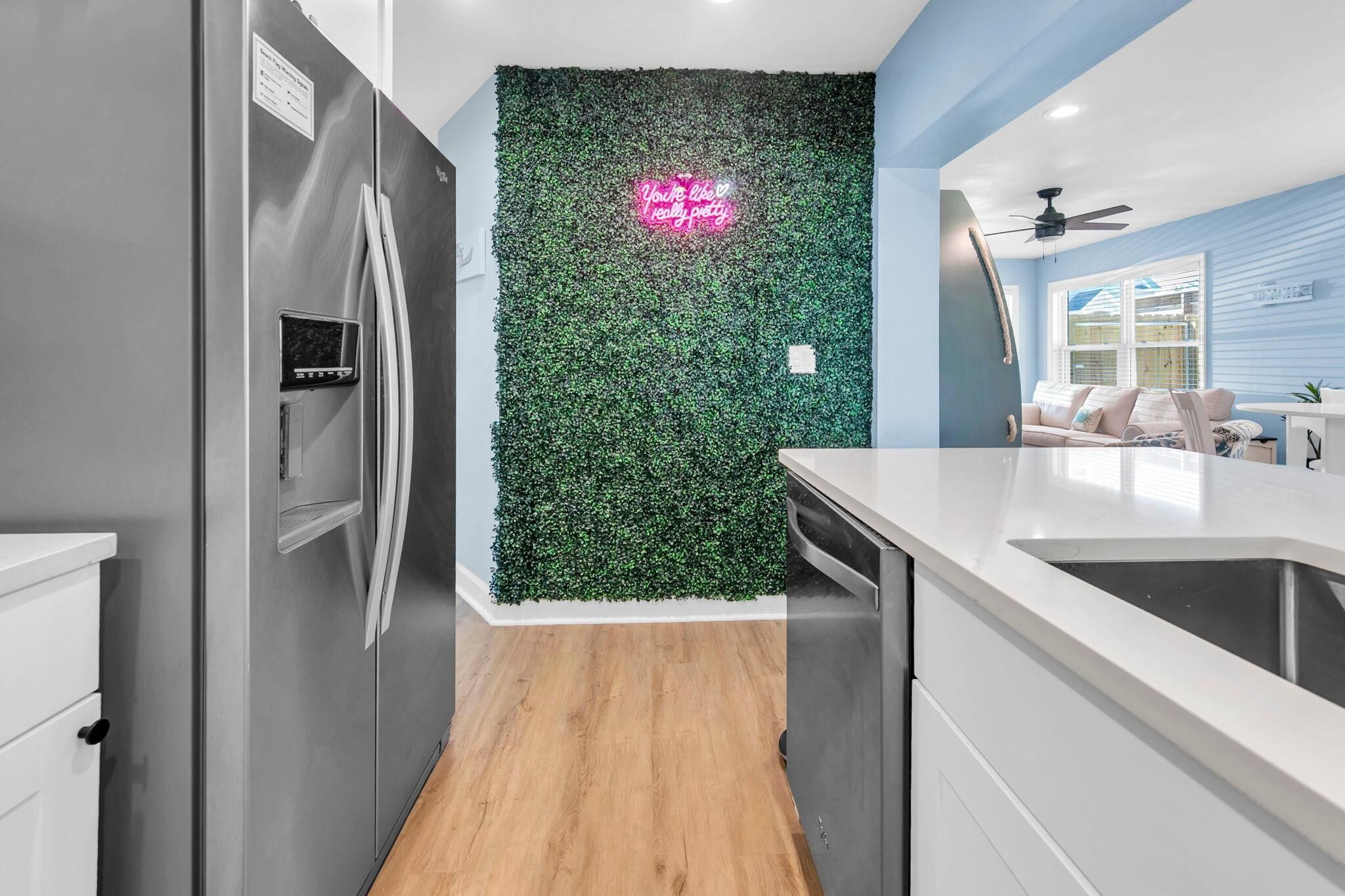 Une cuisine moderne avec un réfrigérateur en acier inoxydable, un plancher en bois, un comptoir blanc et un mur végétal vert.