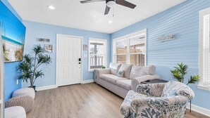 Una sala de estar con un sofá color beige, un sillón estampado, un ventilador de techo y un cuadro grande en la pared.