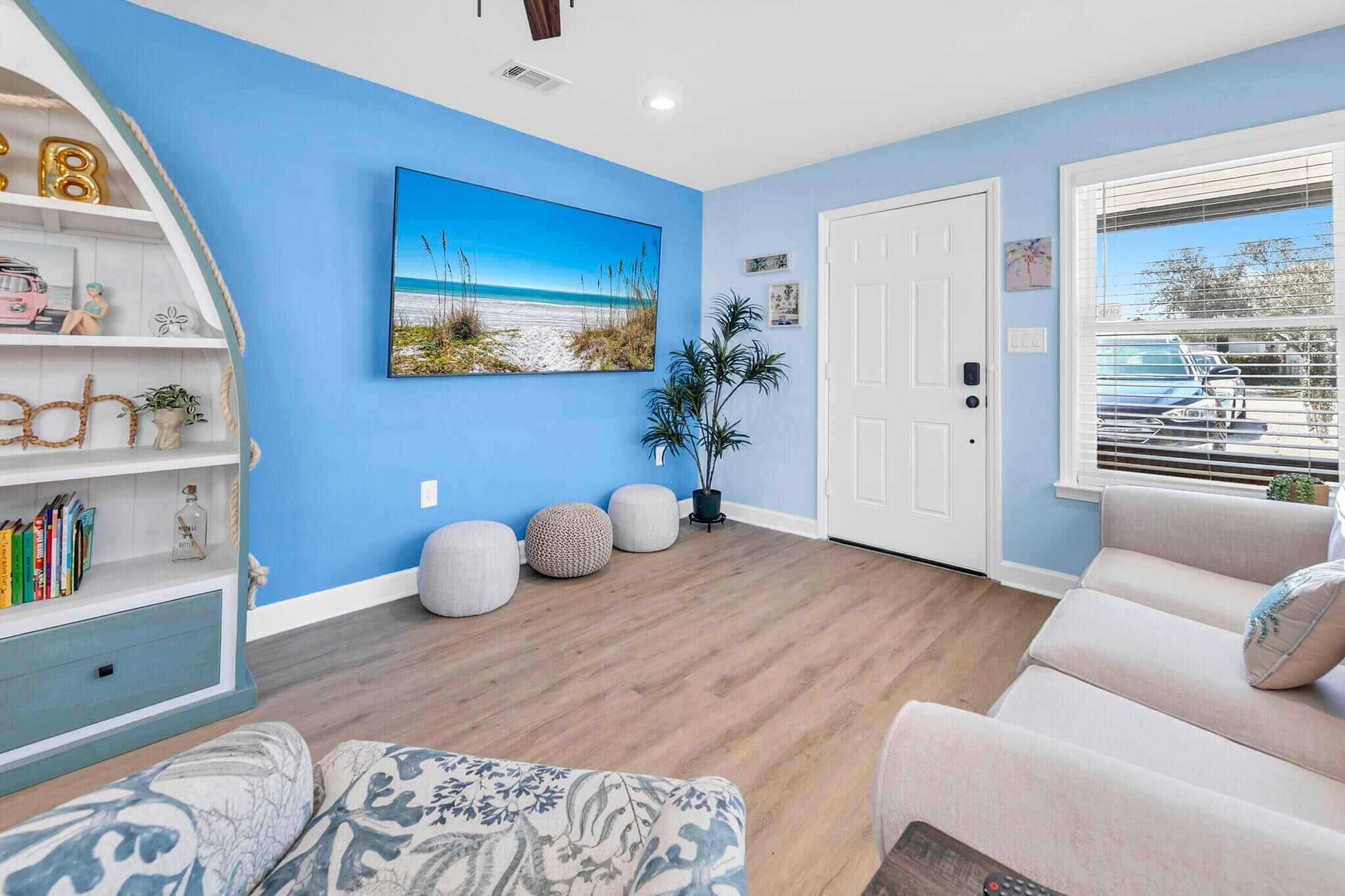 A living room with a blue wall, a beach-themed painting, a white door, and a window with blinds.