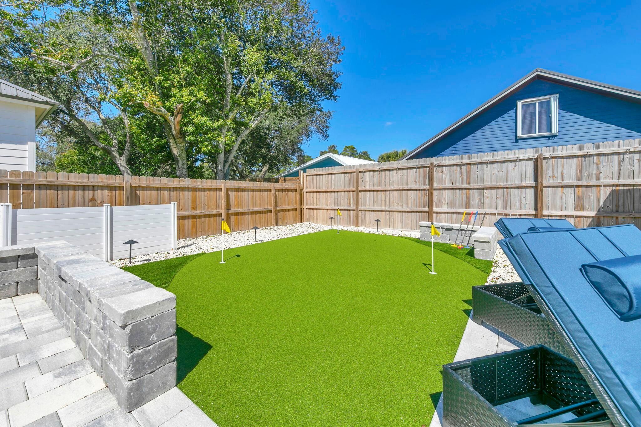 Un jardin avec une pelouse artificielle, un barbecue, une clôture en bois et une maison avec un toit bleu.