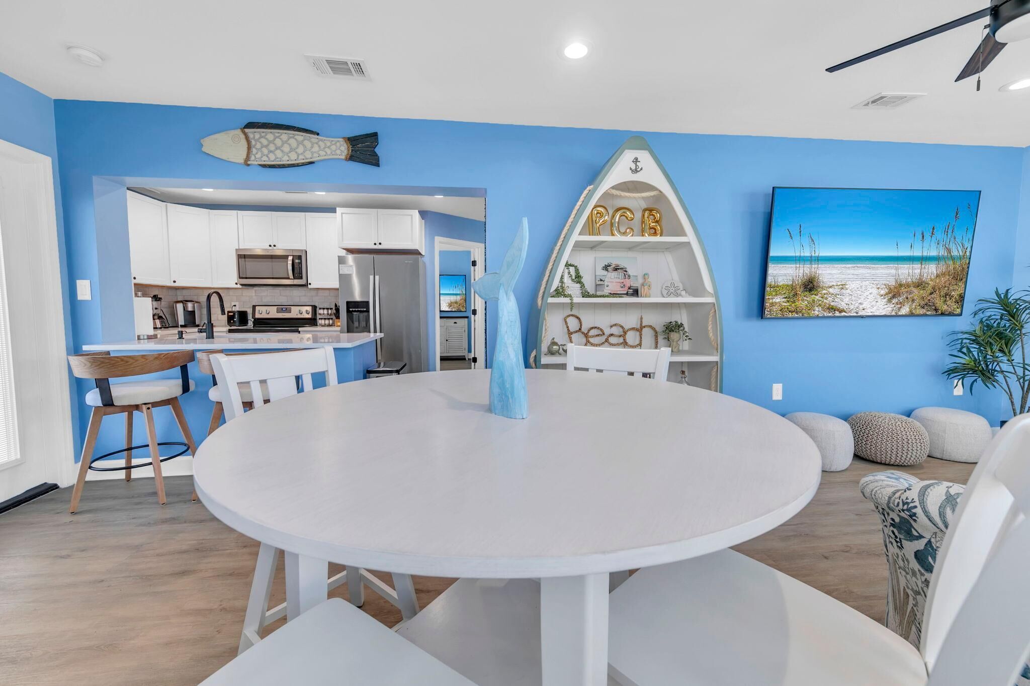 A modern kitchen with white cabinets and a microwave, a dining area with a round table and chairs, and a blue wall with a beach-themed painting and decorative items.