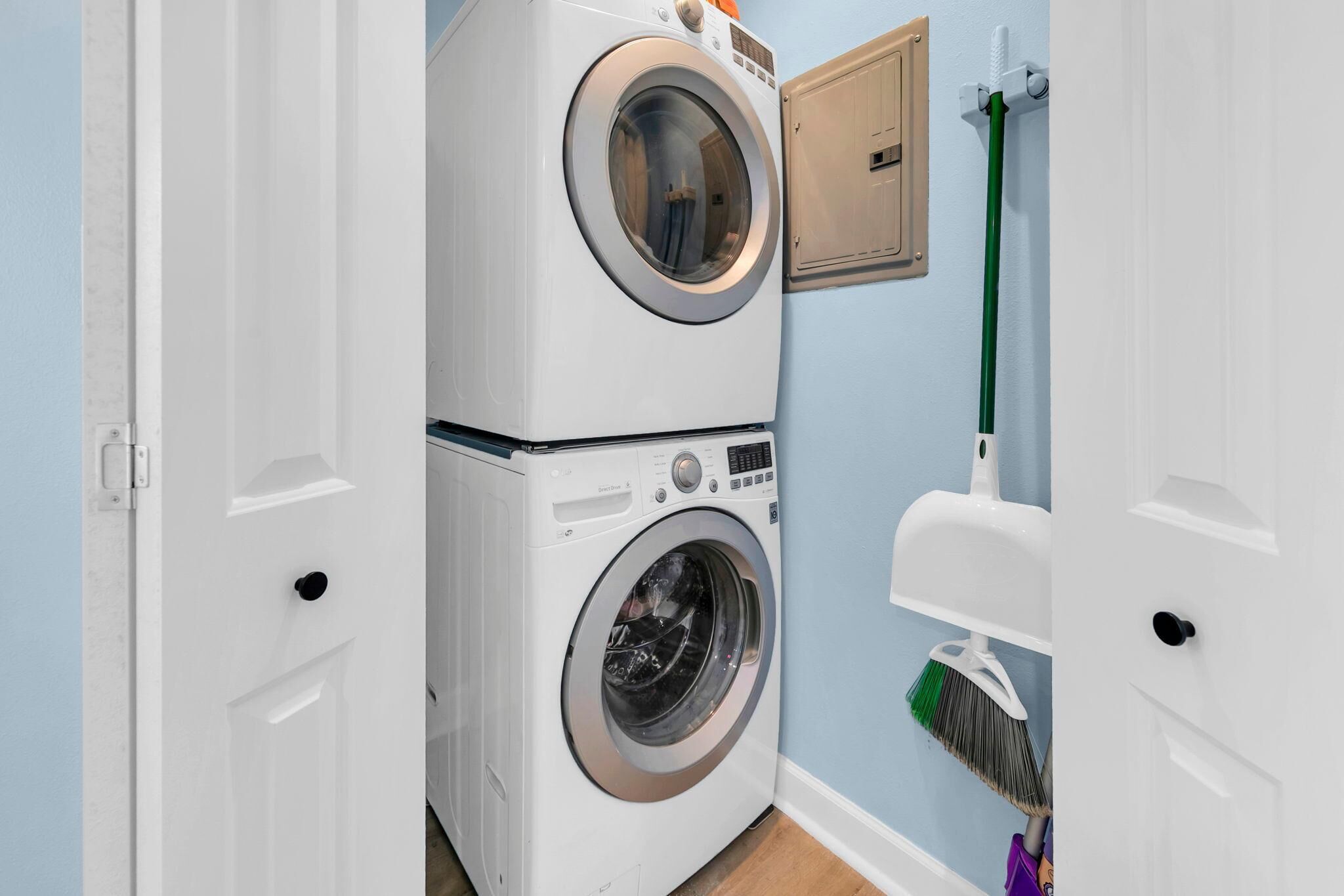 A laundry room with two stacked washing machines, a broom, and a wall-mounted vent.