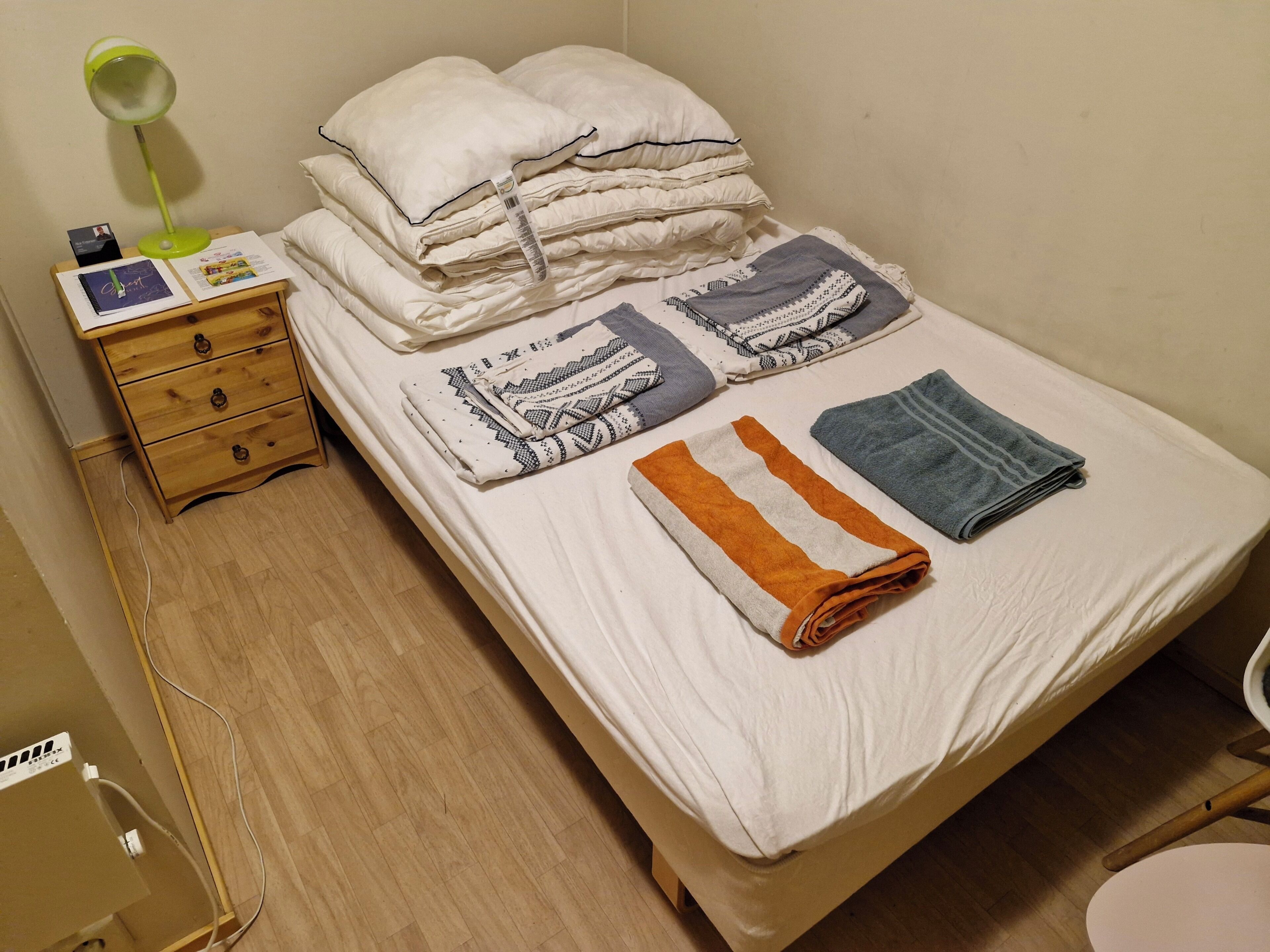 Ein ordentlich gemachtes Bett mit Kissen, gefalteten Decken und Handtüchern. Ein Nachttisch mit Lampe und einigen Gegenständen steht daneben. Der Raum verfügt über Holzböden und eine schlichte Wand.