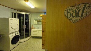 A compact bathroom with a washing machine, a glass shower enclosure, and a wooden door with a 'Bad' sign. - Studio with fitness room (Troms)