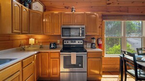 A cabin kitchen with wooden cabinets, a stainless steel oven, a microwave, and a window overlooking greenery. - Ridgecrest Retreat | Hot Tub • Decks • Game Room! (Gatlinburg)