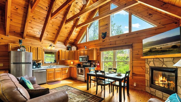 A log cabin interior with a kitchen, dining area, fireplace, and a large window. - Ridgecrest Retreat | Hot Tub • Decks • Game Room! (Gatlinburg)