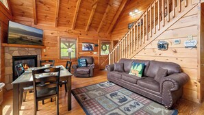 A wooden cabin interior with a fireplace, a dining table with chairs, a leather couch, and a staircase. - Ridgecrest Retreat | Hot Tub • Decks • Game Room! (Gatlinburg)