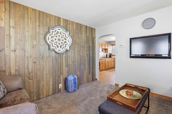 A living room with a wooden wall, a TV, a clock, and a kitchen in the background.