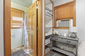 A room with a wooden door, a shower, a vanity with a mirror, a radiator, and a bedside table with a lamp.