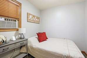 A bedroom with a bed, a bedside table with a lamp, a wall-mounted air conditioner, and a sign that says 'Relax.'