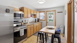 A compact kitchen with wooden cabinets, stainless steel appliances, a small dining area, and a window with a view. - The Appleton Downtown Den (Appleton)