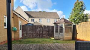 Un jardin avec une terrasse en bois, un petit abri de jardin et une maison en arrière-plan.