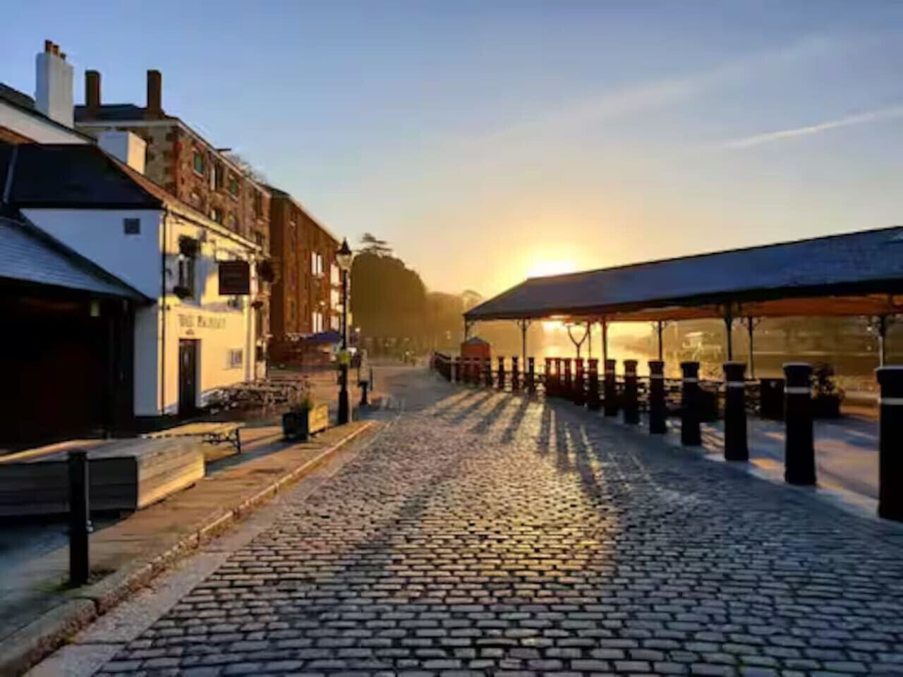 A cobblestone street with buildings on either side, a covered walkway, and a sunset in the background.