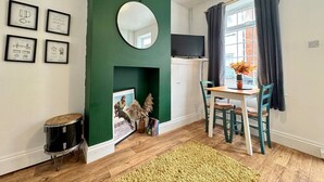 Una habitación con una pared verde de acento, un espejo redondo, cuadros enmarcados, un tambor, una chimenea, una mesa con sillas y una ventana con cortinas.